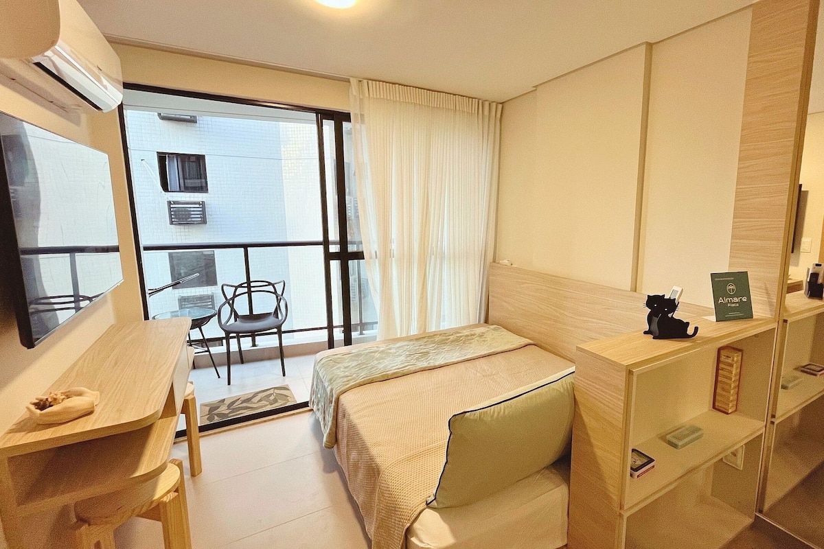 A hotel room with a bed, a desk, a chair, a small table, a mirror, and a view of the outside balcony.
