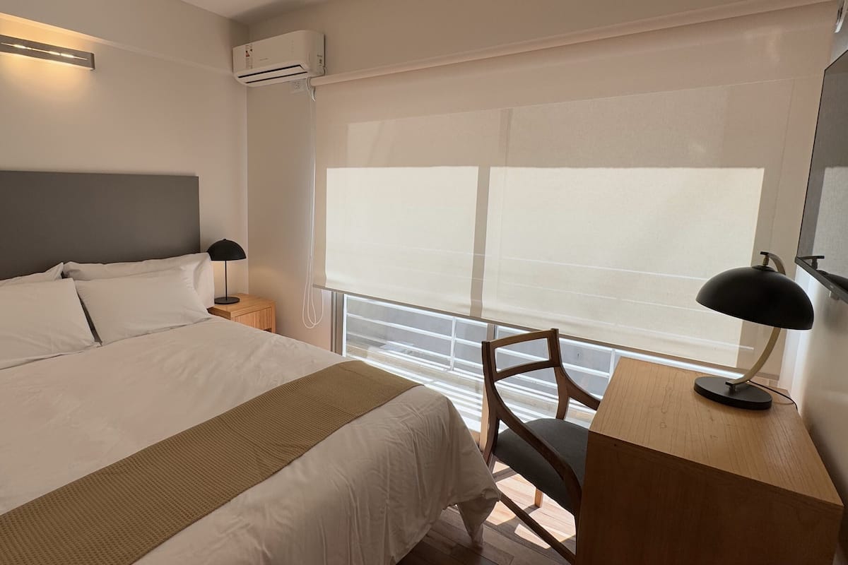 A hotel room with a large bed, a desk with a chair, a lamp, and a window with blinds.
