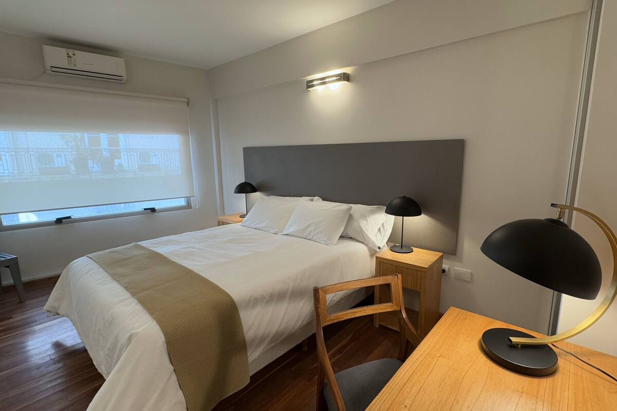 A hotel room with a bed, bedside tables, a desk, a chair, a lamp, a window with a view, and an air conditioning unit.