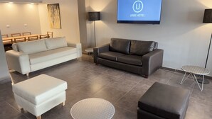 A modern hotel room with a white sofa, a black leather sofa, a small round table, and a flat-screen TV displaying the URBE HOTEL logo. - Urbe Hotel (Buenos Aires)