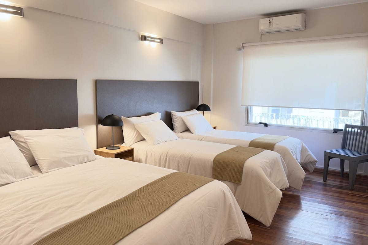 A hotel room with two beds, a window with a view, a chair, and a wall-mounted air conditioner.