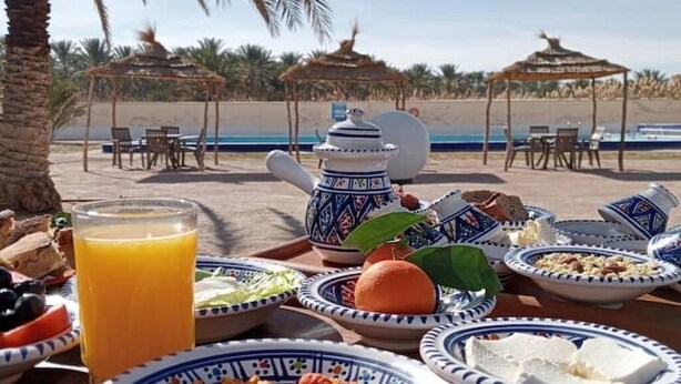 Un desayuno en la playa con una mesa, platos, una tetera y un vaso de zumo de naranja.