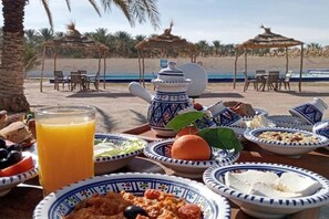 A breakfast setup on a beach with a table, plates, a teapot, and a glass of orange juice.