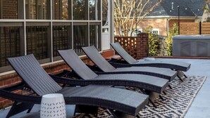 A row of wicker lounge chairs with a patterned rug, a white vase, and a brick wall in the background.