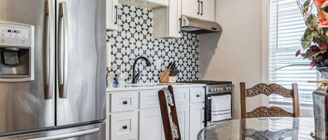 A modern kitchen with a stainless steel refrigerator, white cabinets, a patterned backsplash, and a glass dining table.