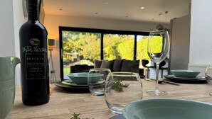 Una mesa de comedor con una botella de vino, copas y platos, en una habitación moderna con un ventanal grande y un sofá al fondo.