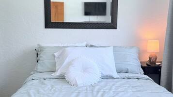 Een opgemaakt bed met witte lakens en een decoratief kussen, een bedlamp en een spiegel aan de muur.