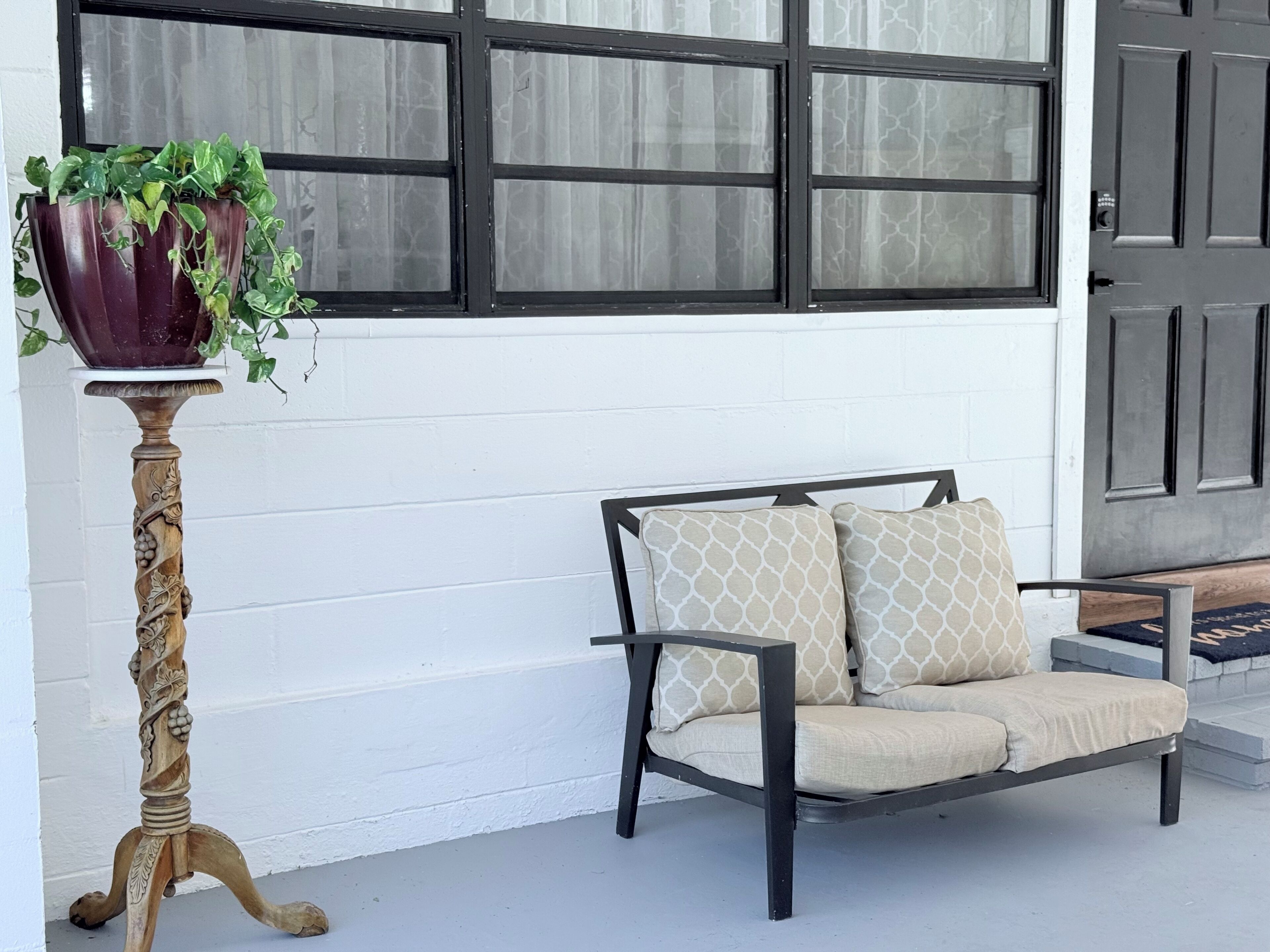 Pared exterior blanca con un ventanal de marco negro, un banco de madera oscura con cojines estampados y una mesa de madera con una planta en maceta.