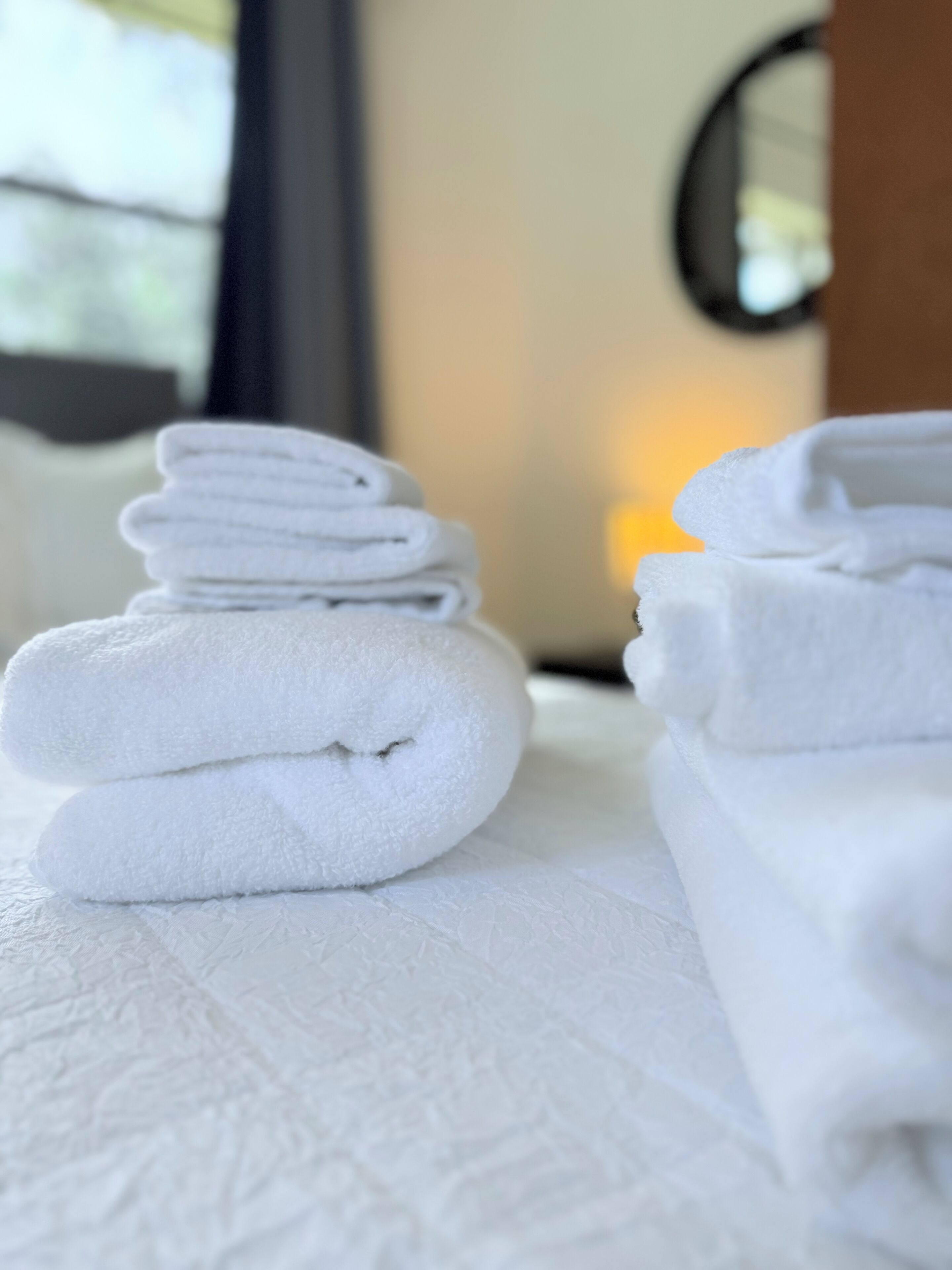A stack of neatly folded white towels on a textured white surface, likely in a hotel room.