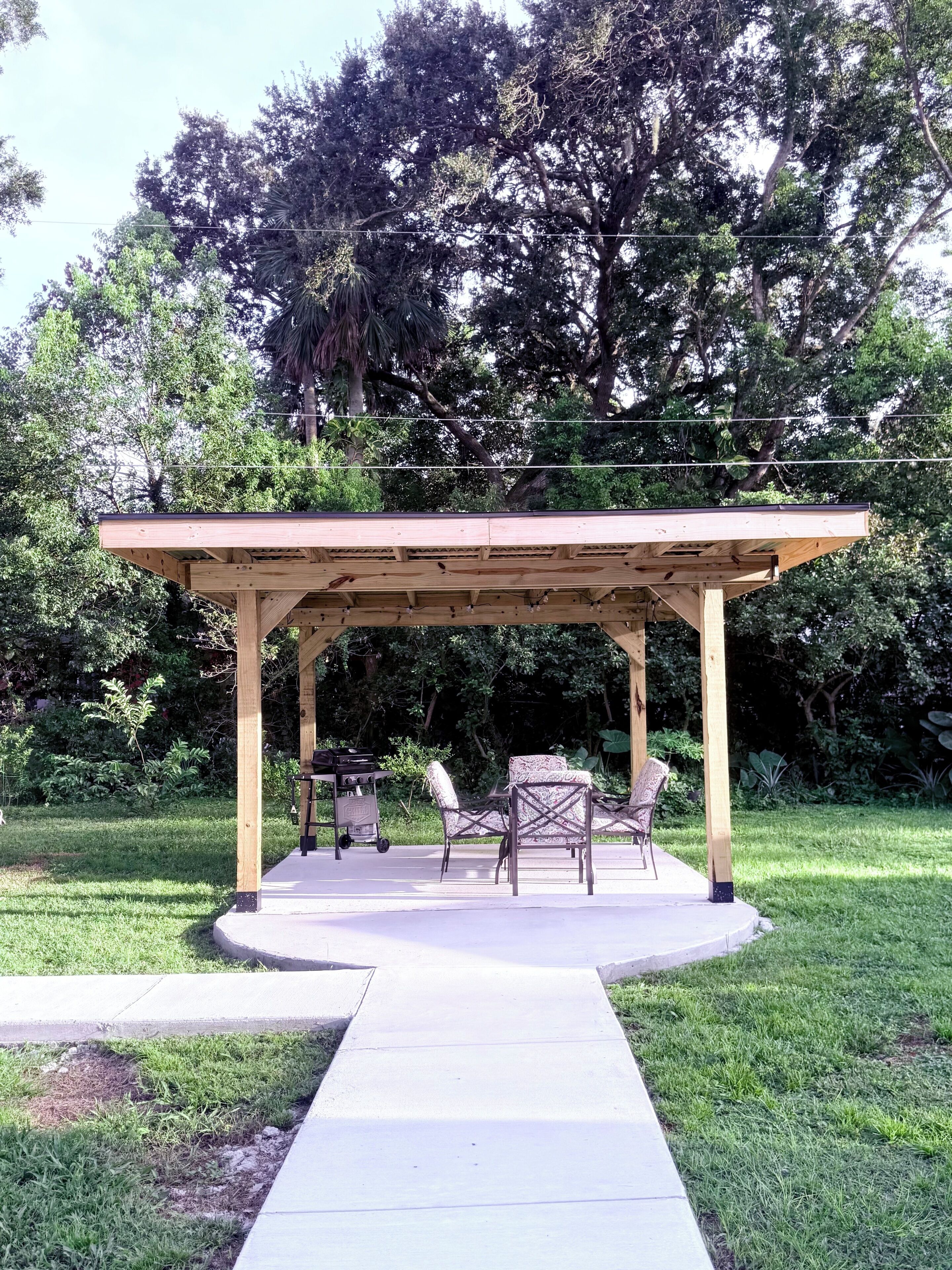 A wooden pergola structure with a concrete pathway leading to it, surrounded by lush greenery and trees.