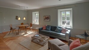 A spacious living room with a grey sofa, wooden coffee table, and a dining area with a wooden table and chairs. - New top-equipped vacation apartment - 20m to the lake in historic monastery fiefdom (Tegernsee)