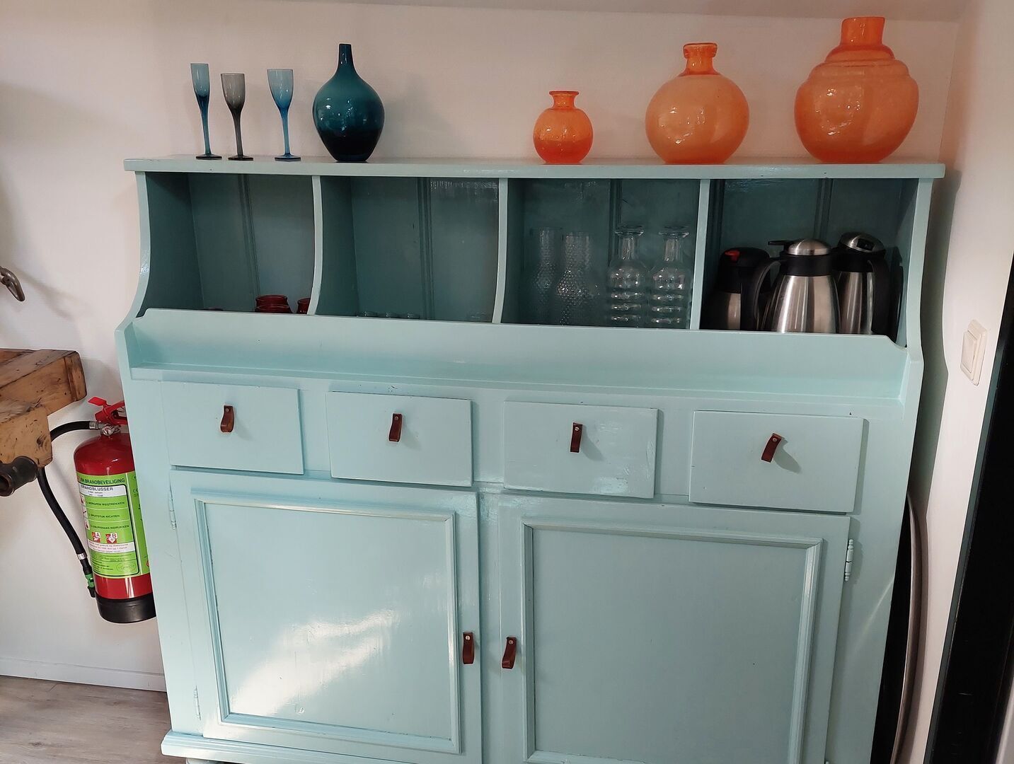 Een lichtblauwe kast met meerdere lades en planken, gevuld met serviesgoed en keukenbenodigdheden.