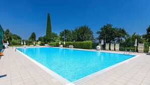 A rectangular swimming pool with a tiled deck, surrounded by white lounge chairs and umbrellas, set against a backdrop of trees and a clear blue sky. - Il Melagrano A12 by Wonderful Italy (Padenghe sul Garda)