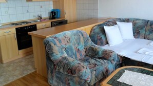 A compact kitchen with wooden cabinets, a built-in oven, and a coffee maker. A sofa with a floral pattern is positioned near the kitchen, and a small table with a newspaper is visible. - Cozy Retreats in Crikvenica Quiet Neighborhood (Crikvenica)