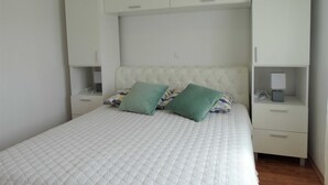 A bedroom with a bed, two pillows, a nightstand, a drawer, and a wardrobe.