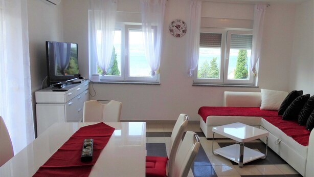 A room with a TV, a dining table, a sofa, and two windows with curtains.