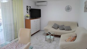 Un salon avec un canapé, un téléviseur, une horloge et une fenêtre équipée de stores.