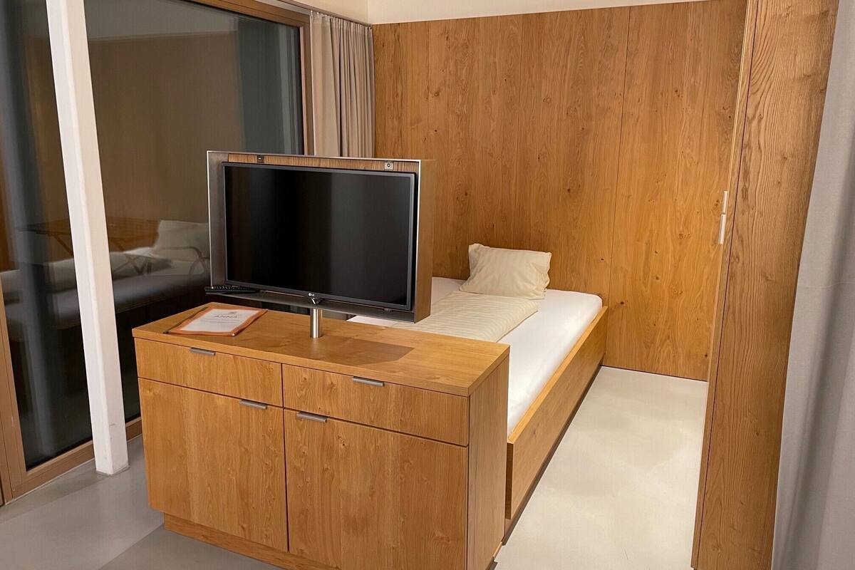 A modern hotel room with a wooden wardrobe, a flat-screen TV, a bed with white bedding, and a large window with curtains.