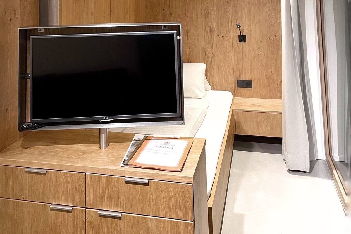 A modern hotel room with a wooden panel wall, a flat-screen TV on a wooden cabinet, a bed with white bedding, and a sliding glass door to the right.