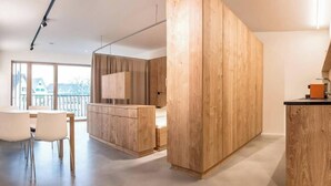 A modern kitchen with wooden cabinets, a dining area with white chairs, and a large window with a view of buildings. - Apartmenthaus Anna - Self-Check-in (Lustenau)