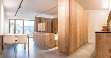 A modern kitchen with wooden cabinets, a dining area with white chairs, and a large window with a view of buildings.