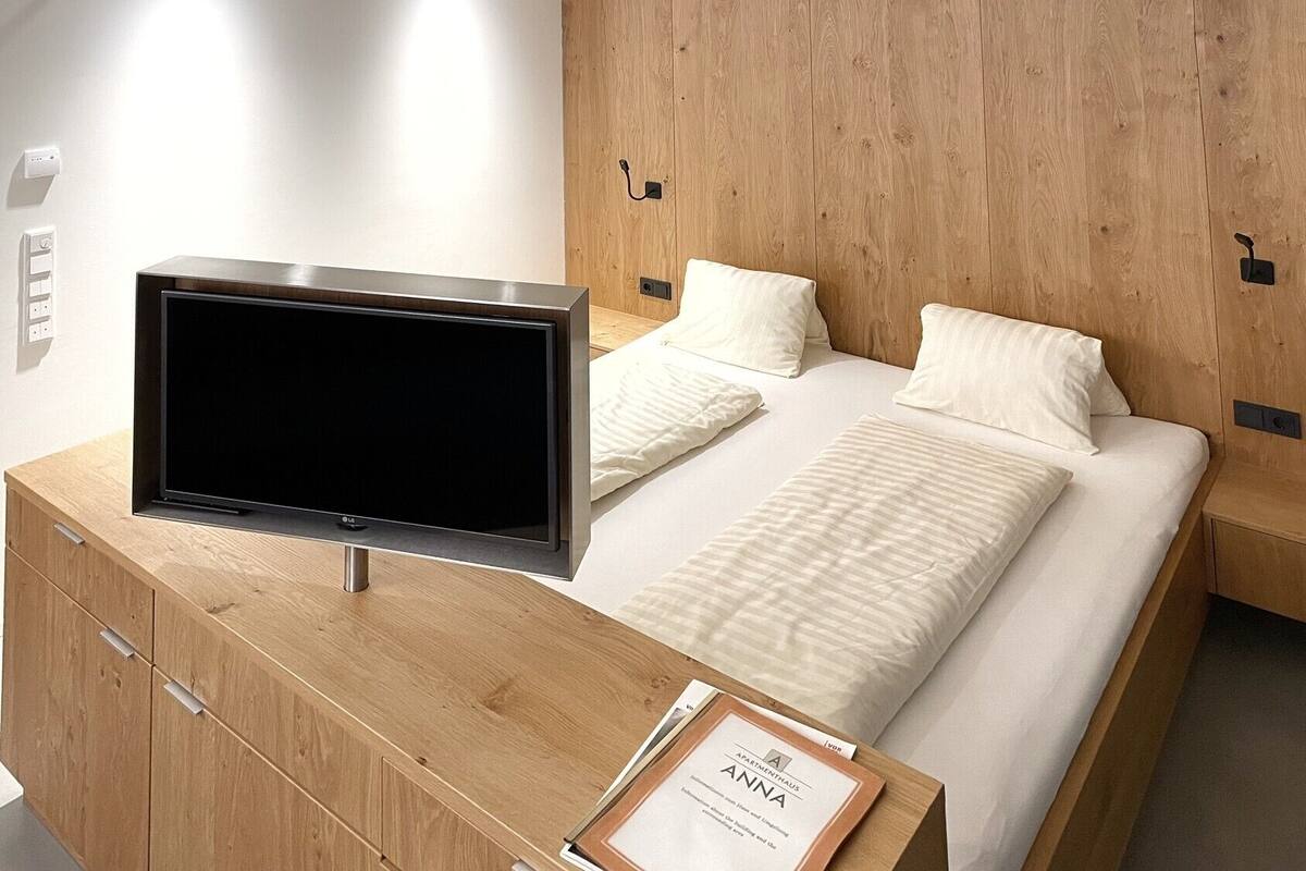 A modern bedroom with a wooden headboard, a flat-screen TV on a wooden dresser, a neatly made bed with white linens, and a framed certificate on the dresser.