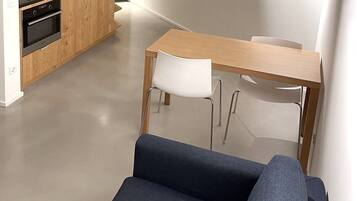 Et moderne køkken med træskabe og en sort bordplade, et spiseområde med et træbord og hvide stole, samt en mørkeblå sofa i forgrunden.