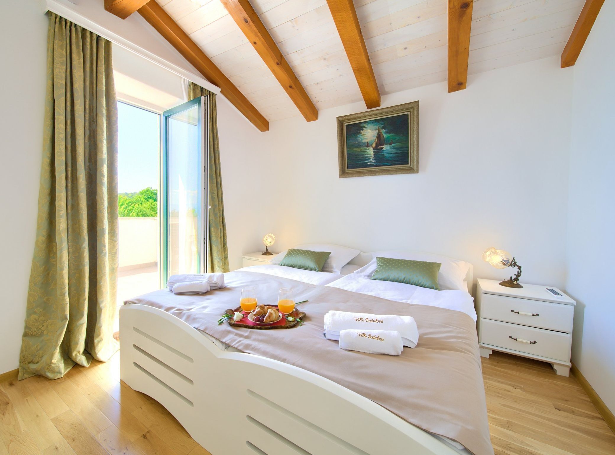 A bedroom with a bed, two bedside tables, a nightstand, a painting, and a window with curtains.