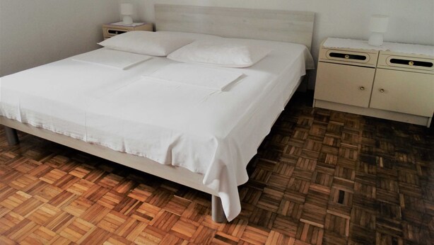 A neatly made bed with white linens, a wooden headboard, and a nightstand with a lamp. The room has a wooden parquet floor. - Airy Crikvenica Apartment With Terrace Close to Beach (Crikvenica)