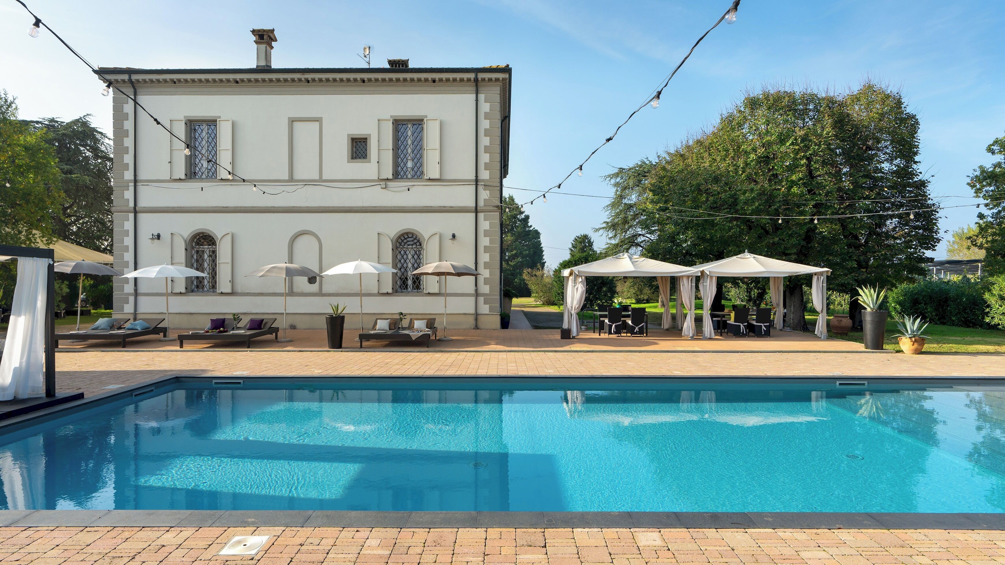 A pool area with lounge chairs and umbrellas, a building with arched windows, and a clear sky.