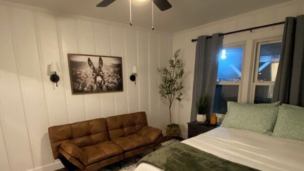 Una camera da letto con un letto, un divano marrone, un tappeto bianco, un quadro appeso al muro e una finestra con tende.