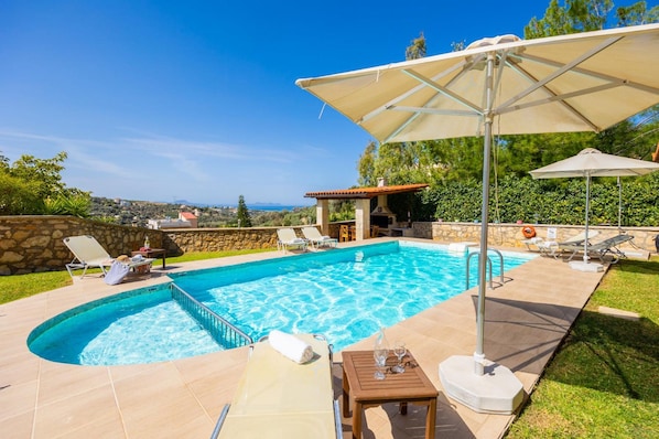 A swimming pool with lounge chairs and umbrellas, surrounded by a stone wall and greenery. - Villa Katia Faragi in Crete (Rethymno)