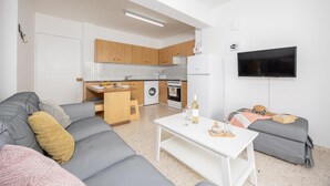 A modern living room with a grey sofa, a white coffee table, and a grey couch. There is a kitchen area with wooden cabinets and a built-in oven. A flat-screen TV is mounted on the wall. - GE Harbour Apartments 16 (Polis)