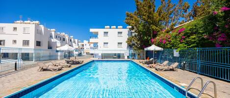 A swimming pool with lounge chairs and a stone deck, surrounded by white buildings and trees with pink flowers.