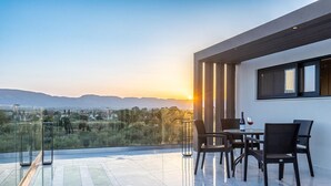 A modern outdoor terrace with a glass balustrade, a dining area with chairs and a table, and a view of the landscape during sunset.