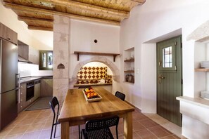 Una cucina con un tavolo di legno, sedie e una ciotola di frutta. Si vede una porta verde e un bancone bianco.
