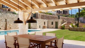 An outdoor patio with a wooden table and chairs, a stone wall, and a pool. - Villa Nafsika in Vamos (Apokoronas)
