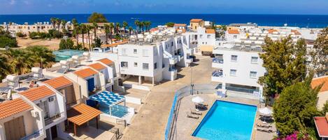 A residential area with a swimming pool surrounded by white buildings with red roofs, a clear blue sky, and the sea in the background.