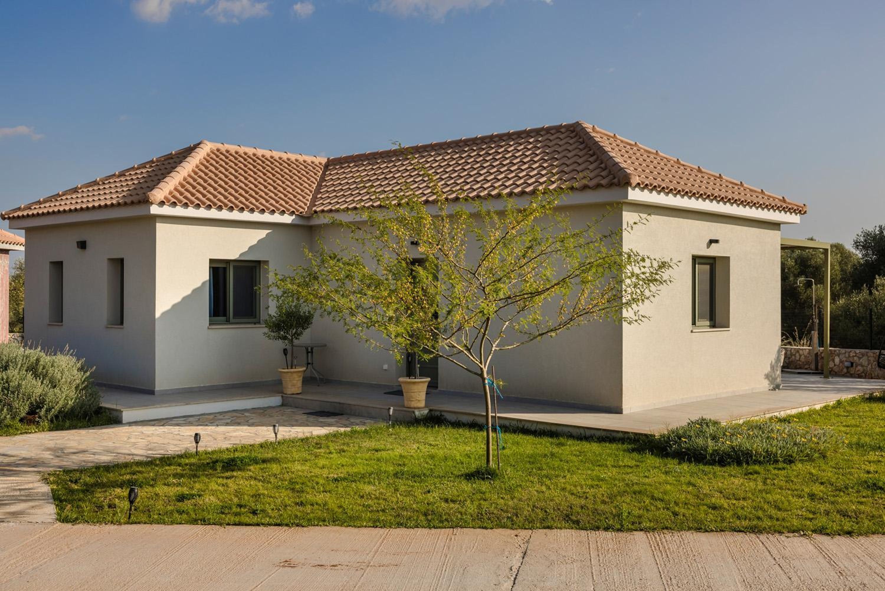 A modern two-story house with a tiled roof, a small front yard with a tree, and a pathway leading to the entrance.