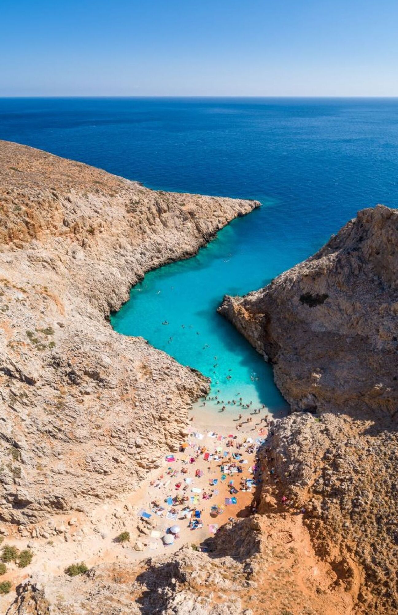A rocky coastline with clear turquoise waters, a sandy beach with umbrellas, and a clear sky.