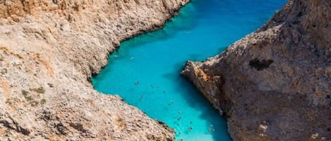 A rocky coastline with clear turquoise waters, a sandy beach with umbrellas, and a clear sky.