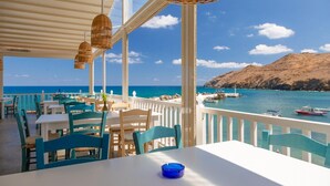 En restaurant ved havet med udendørs siddepladser, hvide borde og blå stole, med udsigt over havet og en stenet kystlinje.