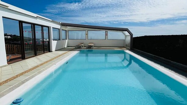 A modern infinity pool with clear blue water, surrounded by a white building with large windows and a glass barrier. - Villa Asomada in La Asomada (Tías)