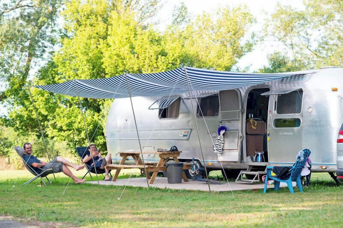 Un camping-car argenté avec un auvent rayé, une table de pique-nique et deux chaises de jardin.