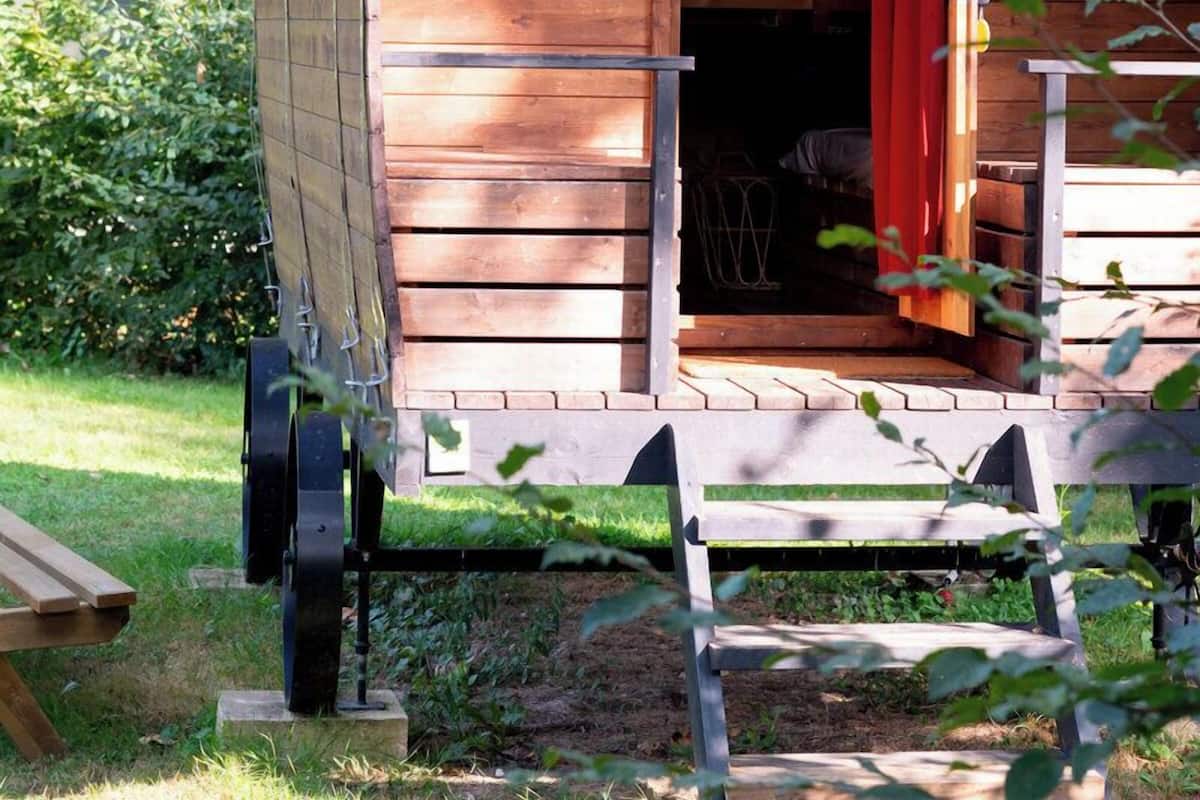 Une structure en bois, abritée et dotée d’une porte ouverte, avec un banc à l’extérieur et une brouette à proximité.
