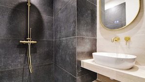 Un baño moderno con una zona de ducha revestida de azulejos negros, un lavamanos blanco con grifo dorado y un espejo con marco dorado.