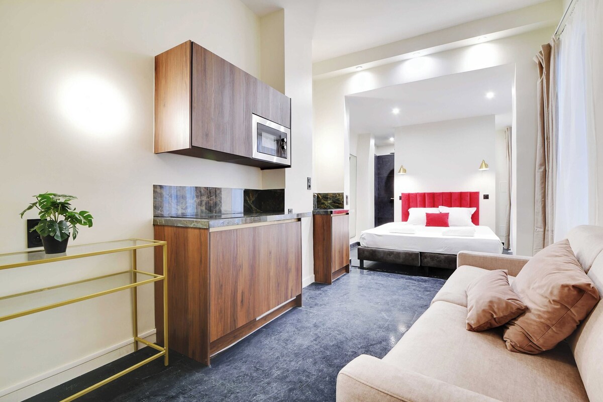 A modern hotel room with a kitchenette, a sofa, and a bed with red and white bedding.