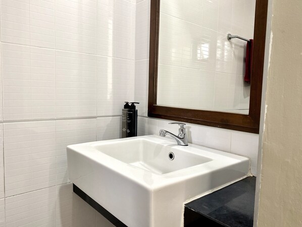 A bathroom with white tiled walls, a white sink with a chrome faucet, and a mirror with a wooden frame. - Mamatel Boutique (Kamala)