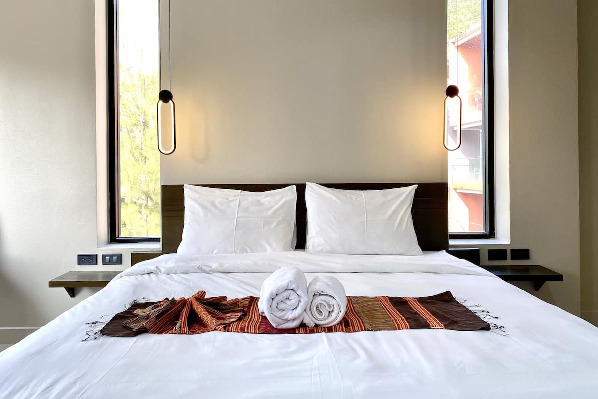 A neatly made bed with white linens, a brown and red patterned throw, and two rolled towels.