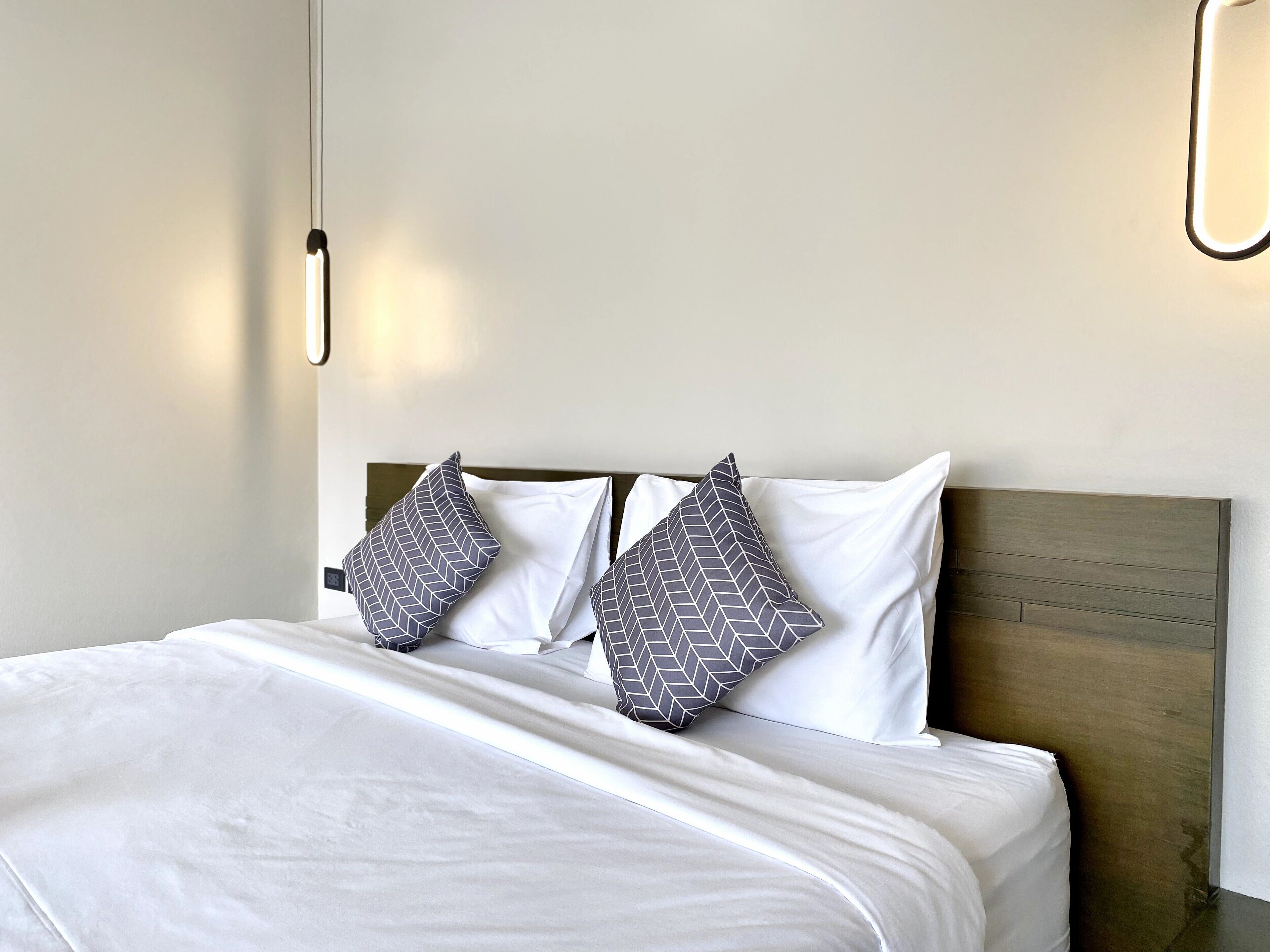 A neatly made bed with white bedding and two patterned pillows, a wooden headboard, and a simple wall with a light fixture.